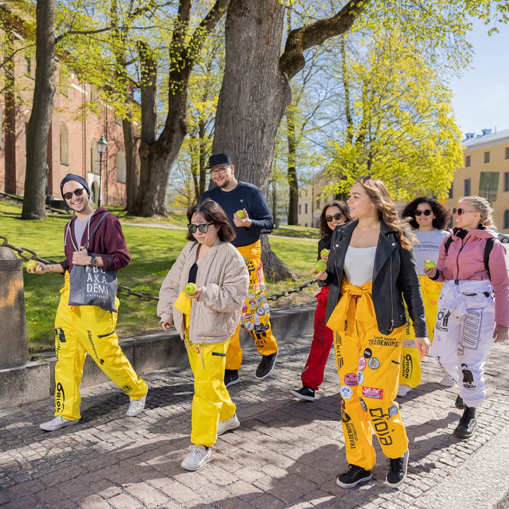 Studenter klädda i overall går med äpplen i händerna i en solig stad.