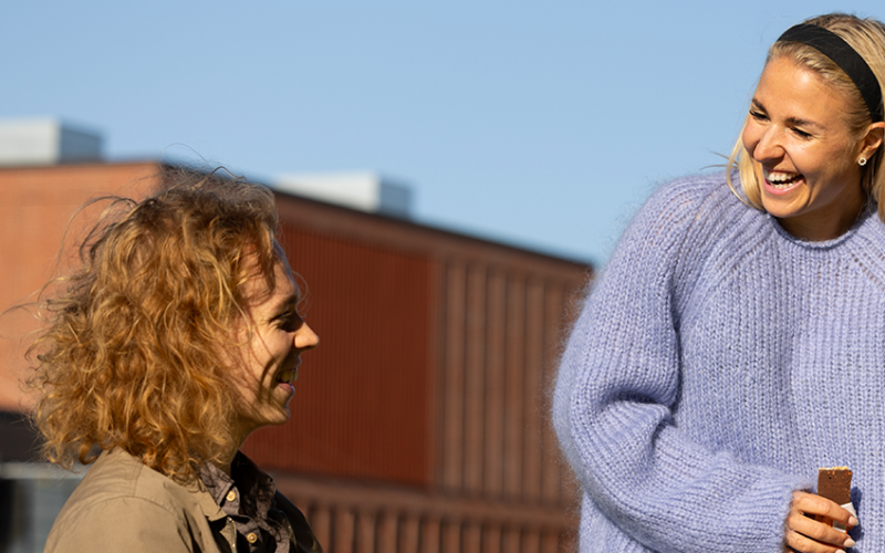 Neljä opiskelijaa hymyilee pihalla auringonpaisteessa. Kolme heistä istuu ja yksi seisoo, taustalla näkyy Otaniemen kampusaluetta.
