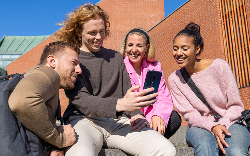 Fyra studenter tillbringar tid på campusgården, ser glada ut och tittar på en mobiltelefon tillsammans.