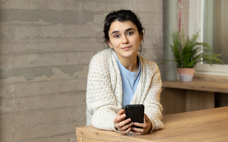 Student leaning against a table with a phone, looking into the camera